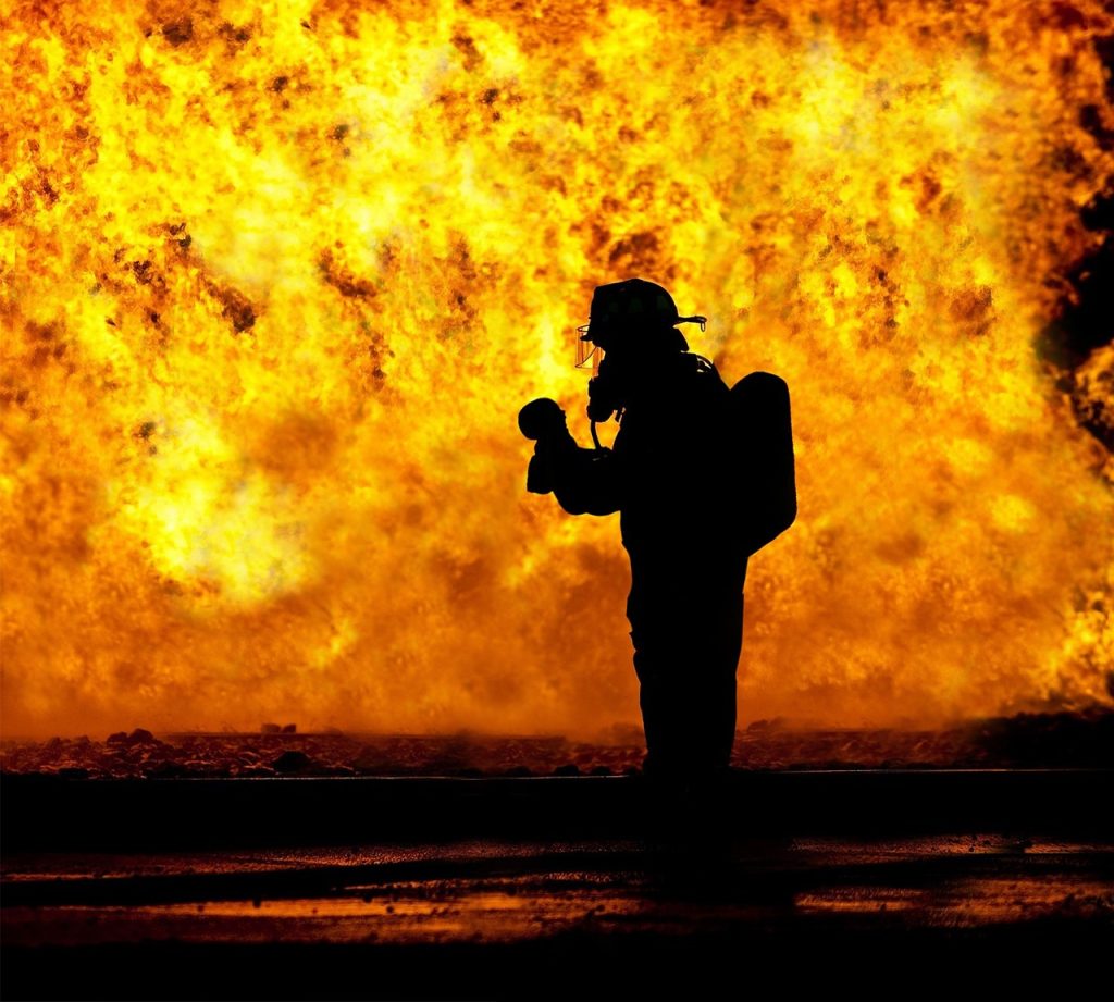 An image showcasing a fire-damaged house engulfed in flames, with firefighters in protective gear battling the blaze amidst billowing smoke, while specialized equipment for fire damage restoration is visible nearby